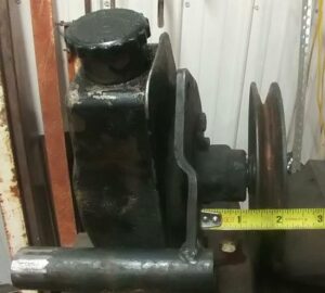 A close-up technical photo showing a Saginaw power steering pump and pulley on a workbench. A yellow tape measure is held against the bracket face, indicating a 1.75-inch measurement to the center of the V-belt groove.