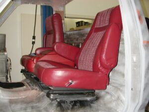 A high-angle interior shot of a white truck cab featuring two freshly upholstered red bucket seats with armrests, mounted on a black metal frame that spans the width of the transmission tunnel.