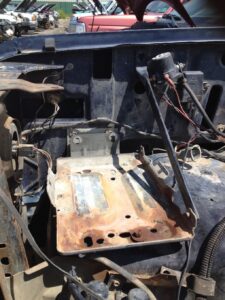 Comparison of original 1980-1986 Ford truck battery hold-down vs later wedge-style tray upgrade.