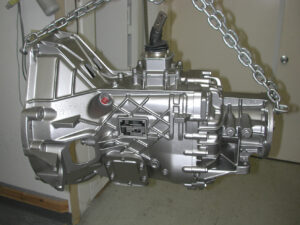 A freshly media-blasted and painted silver aluminum ZF5 transmission casing hanging from a shop crane, showing the integrated bellhousing and shift turret.