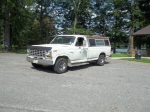 A white 1981 Ford F-150 Custom with a silver camper shell and factory argent wagon wheels, featured as Entrant #16 in the 2020 Virtual Truck Show.